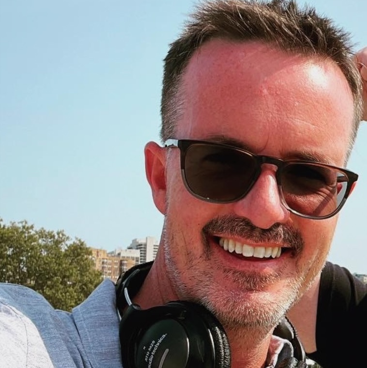 A man smiling outdoors wearing sunglasses and headphones around his neck, with trees and buildings in the background.