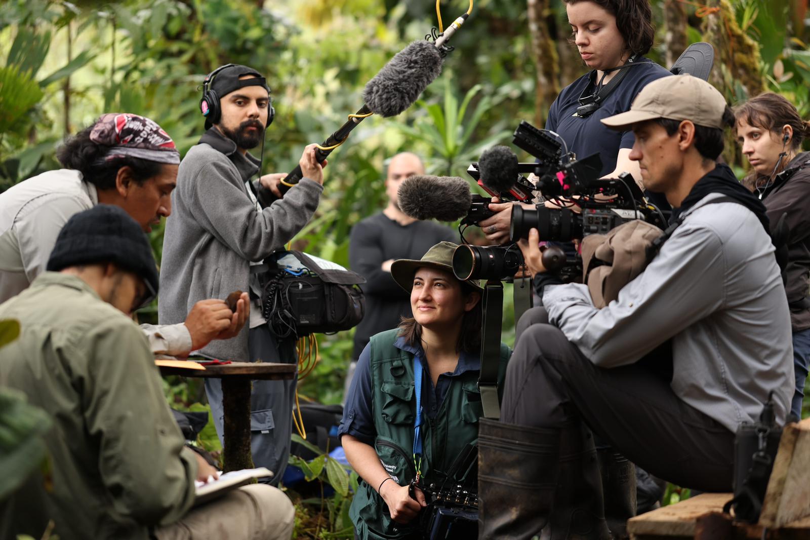 A film crew works together in a dense green forest, capturing footage as a woman in a wide-brimmed hat looks on with a smile.