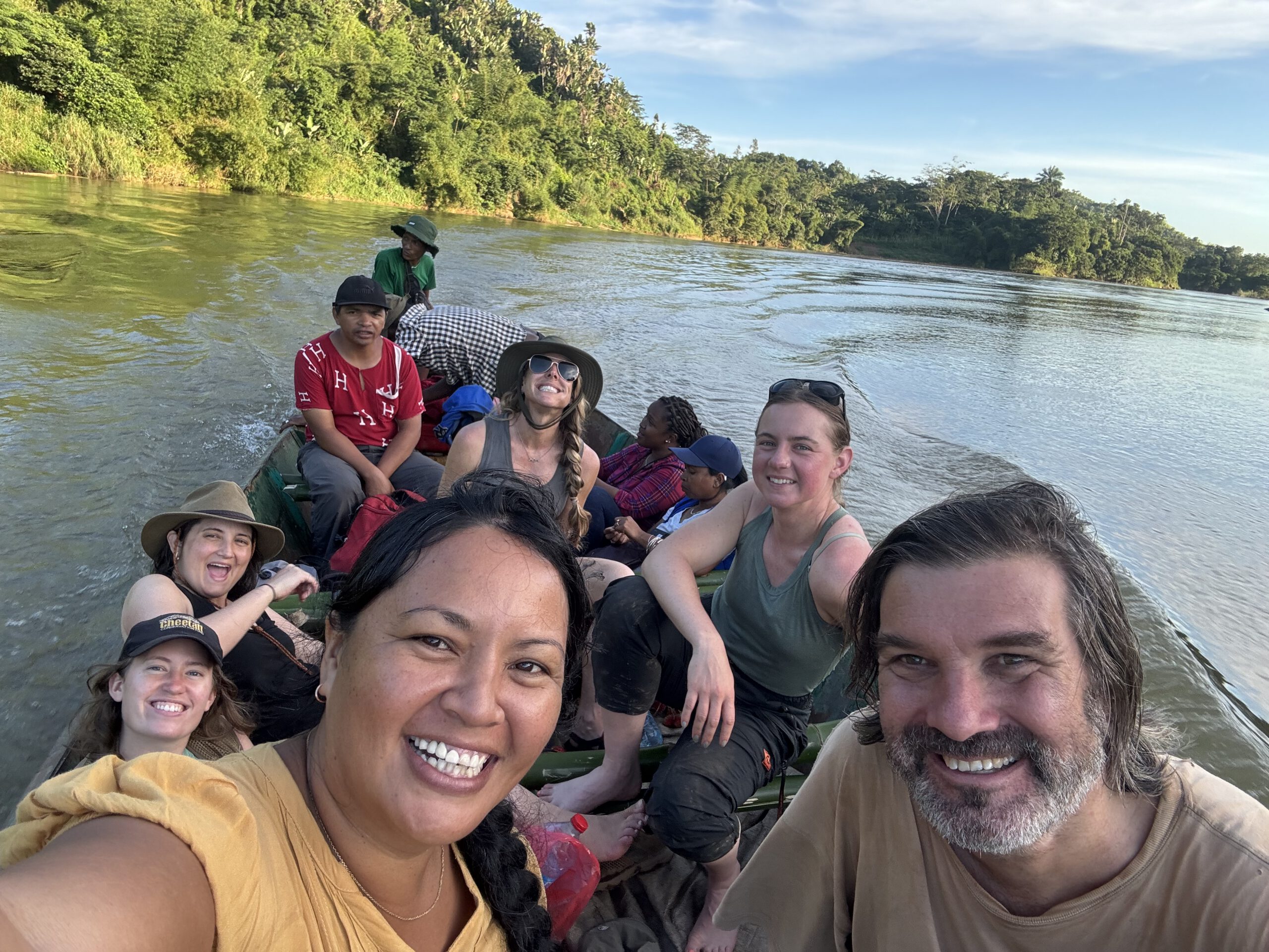 A smiling group of people in a small wooden boat travel down a wide, green river surrounded by lush tropical forest.