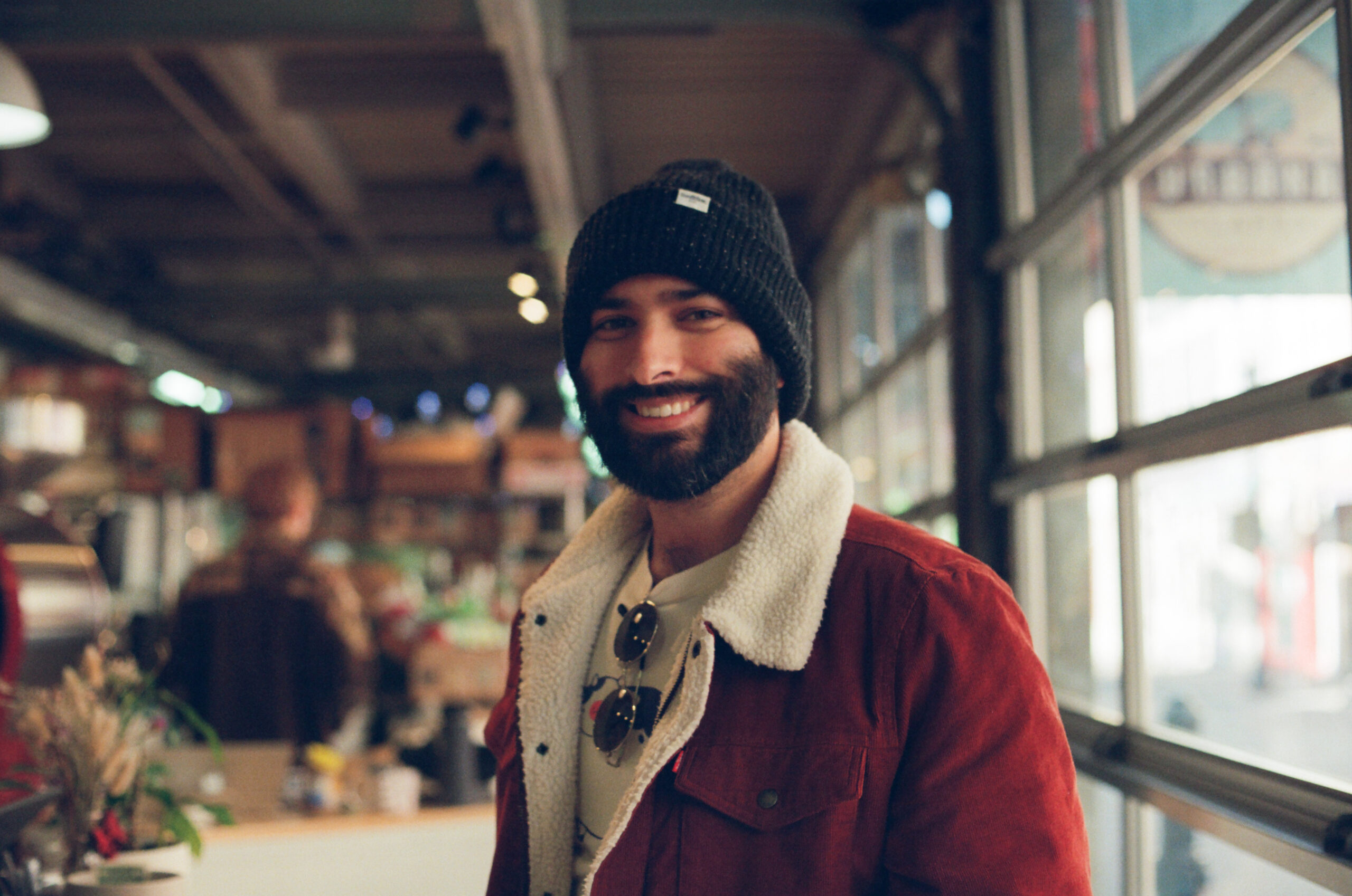 A man in a beanie and red jacket with a shearling collar smiles inside a cozy café with warm light.