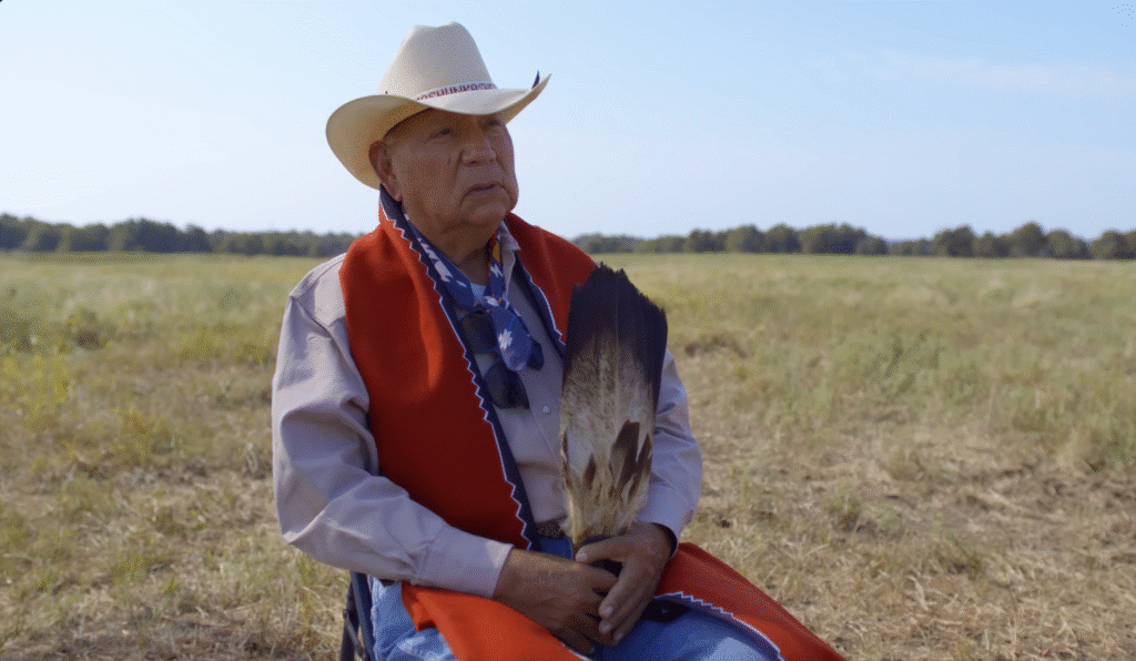 An Indigenous elder wearing a cowboy hat, red shawl, and holding a feather fan sits in a grassy field under a clear blue sky.