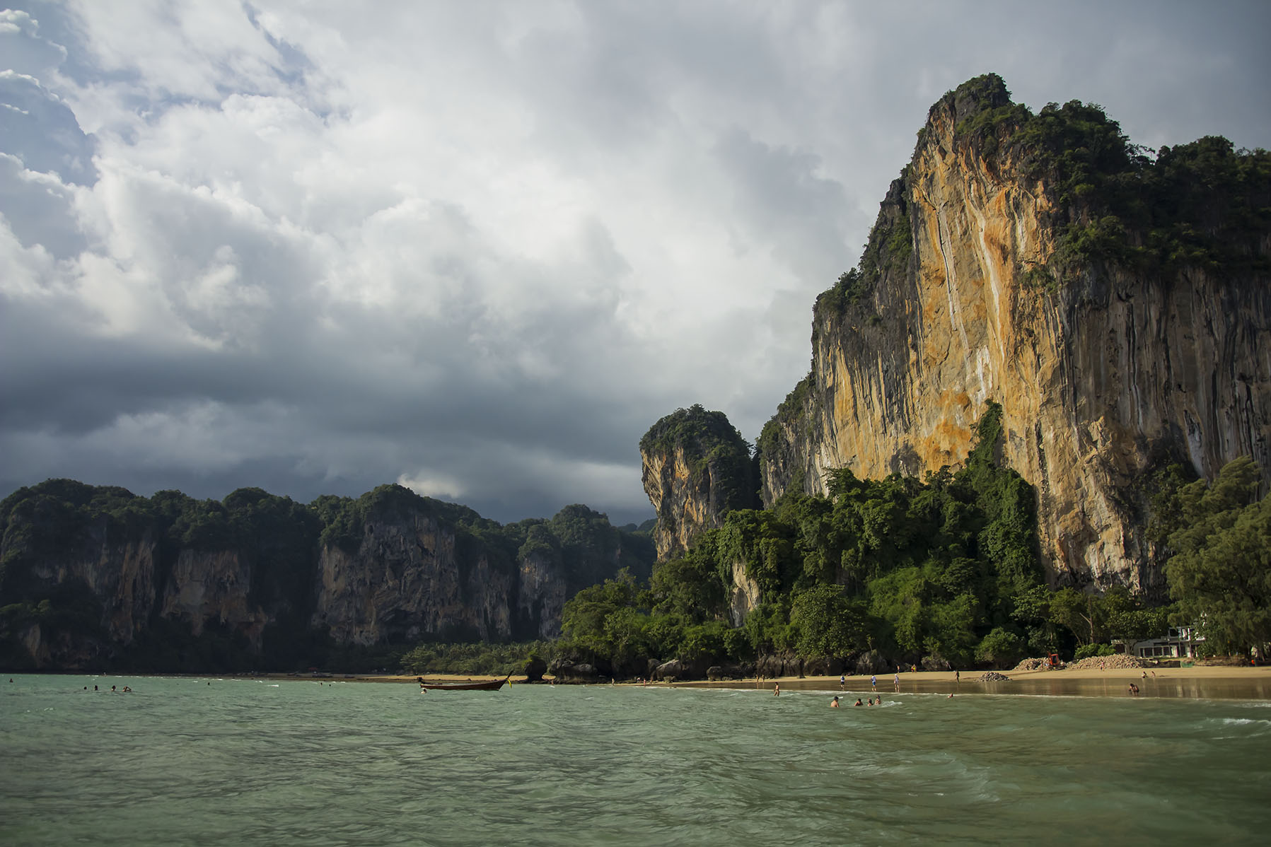 A scenic coastal landscape in Krabi, Thailand, showing tall limestone cliffs rising above turquoise water under dramatic storm clouds.