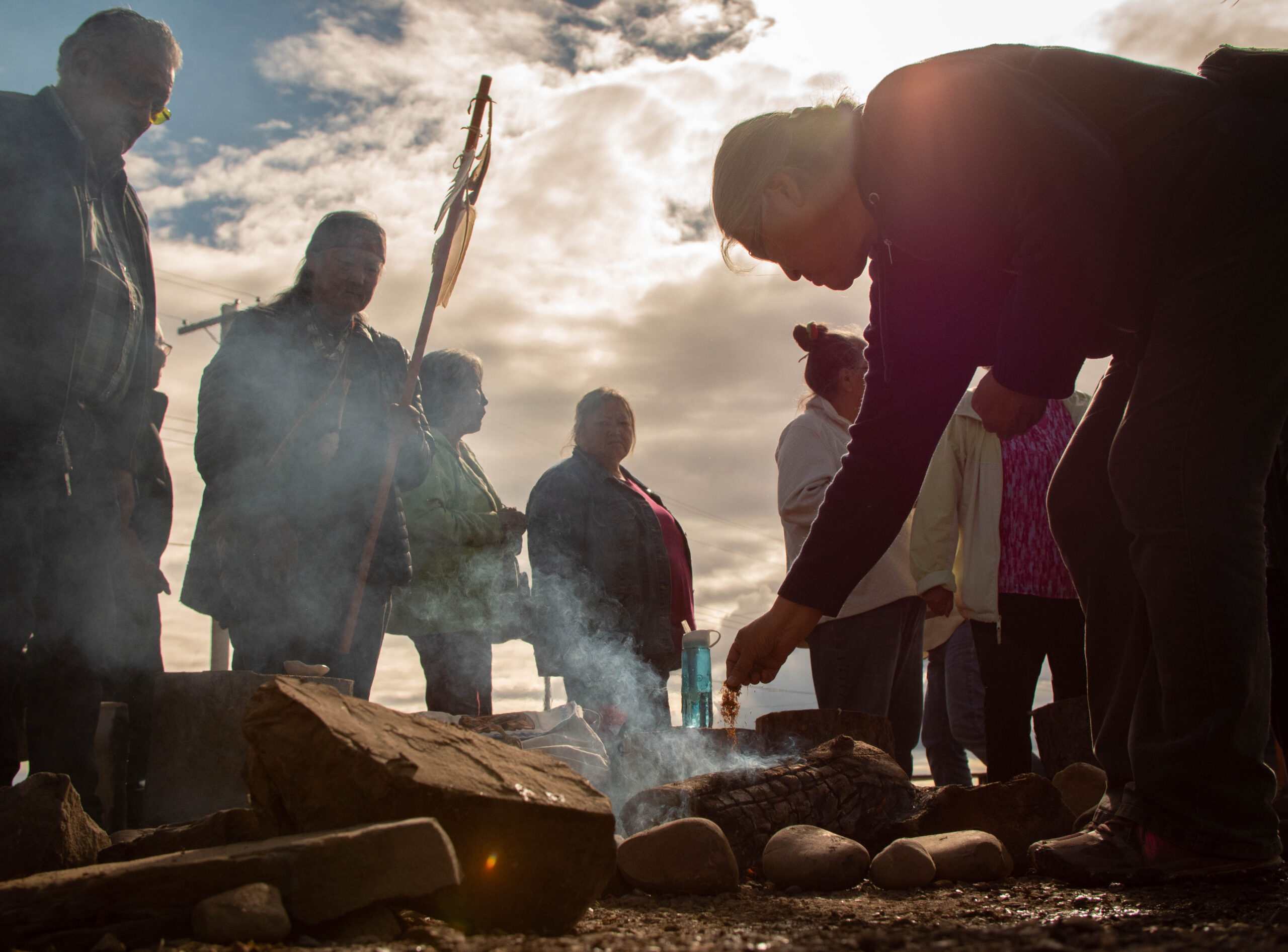 A group gathered around a small fire during a closing ceremony, one person making an offering as smoke rises into the light.