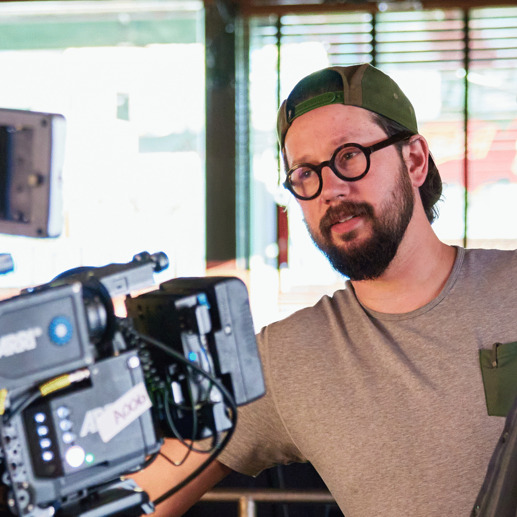 NIck in glasses and a backward cap adjusting a professional video camera indoors under warm light.