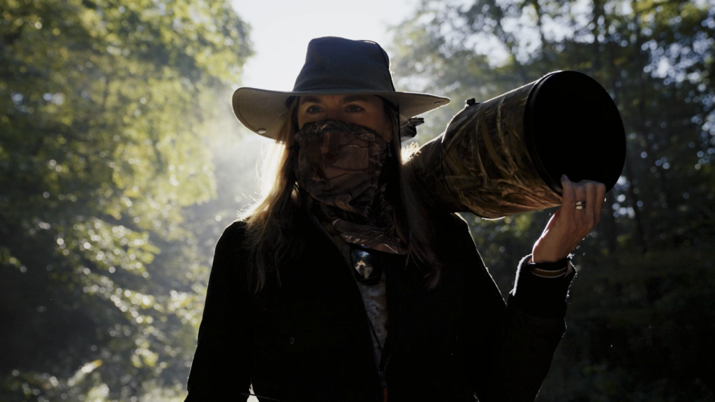 Wildlife photographer standing in a forest holding a large camouflage telephoto camera lens over one shoulder, wearing a wide-brimmed hat and face covering.
